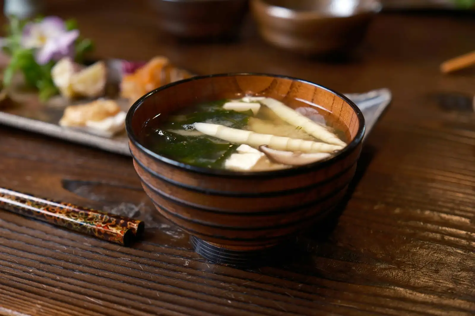 黒い木の卓に置かれた木の椀。澄んだ汁にきのこと青菜